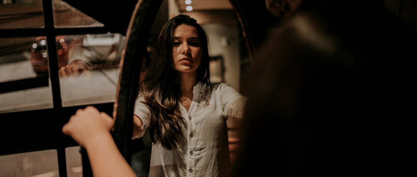 Mulher refletindo no espelho num ambiente à meia luz. A iaem representa o autoconhecimento e a autorregulação das próprias emoções que vêm com a inteligência emocional.